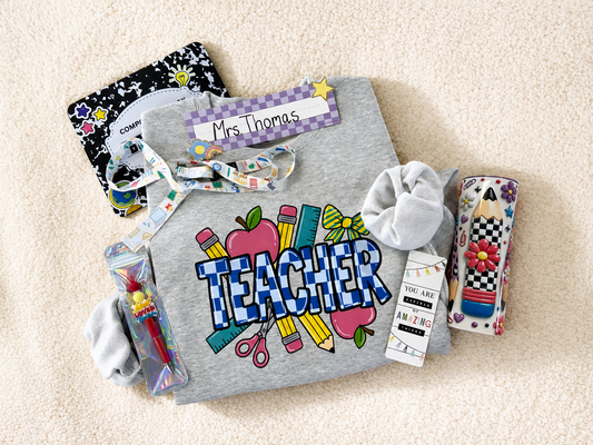 A flat-lay of a Teacher-themed gift bundle on a beige carpet background, including a gray sweatshirt with a colorful 'TEACHER' graphic, a black-and-white star print notebook, a patterned wristlet, a scrunchie, and small accessories.
